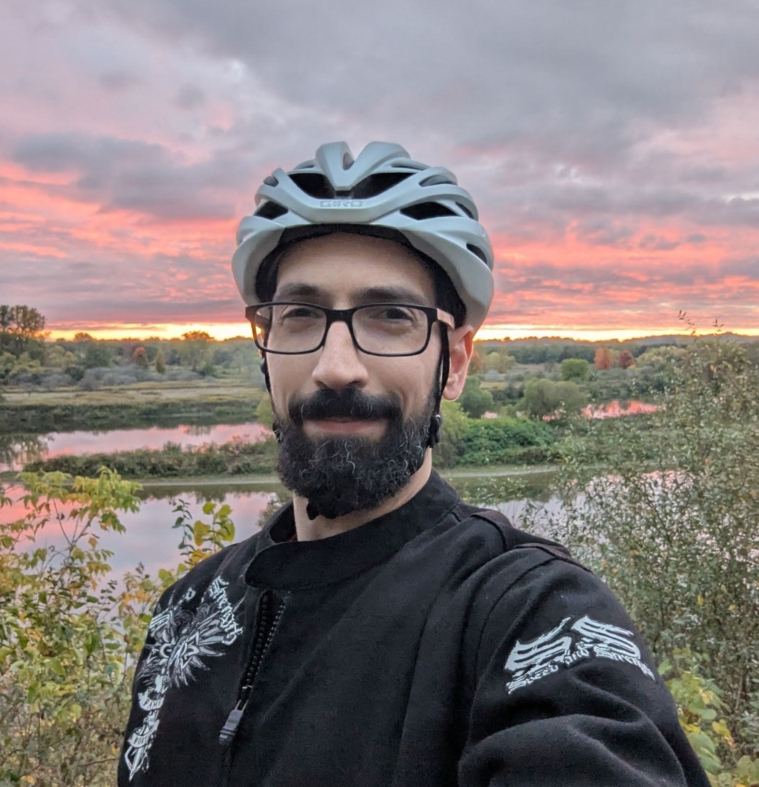 Abdulla Qeblawi smiling outdoors in a bike helmet and glasses, with a river and sunset sky in the background.